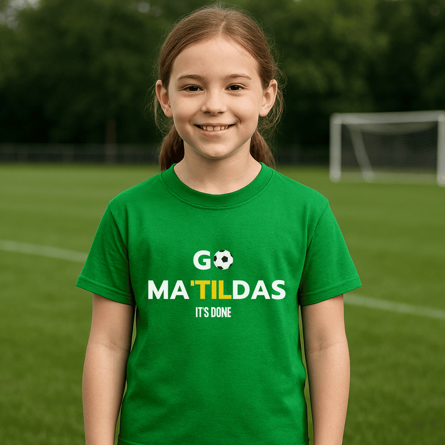 Young girl wearing a green t-shirt with 'GO MATILDAS IT'S DONE' text on a soccer field.
