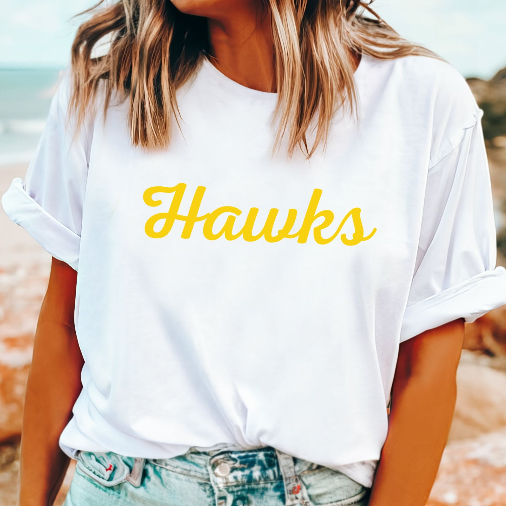 A woman wearing a white short-sleeve Hawthorn Hawks t-shirt with the word 'Hawks' printed in yellow script across the chest, paired with light-wash denim jeans, set against a blurred beach background.