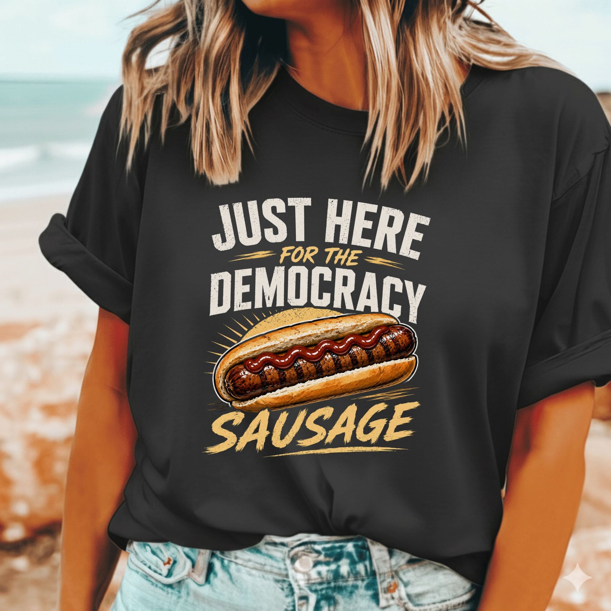 A woman wearing a black graphic t-shirt with a hot dog and the text 'JUST HERE FOR THE DEMOCRACY SAUSAGE' printed on the front, set against a blurred beach background.