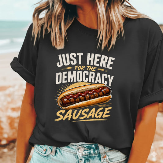 A woman wearing a black graphic t-shirt with a hot dog and the text 'JUST HERE FOR THE DEMOCRACY SAUSAGE' printed on the front, set against a blurred beach background.