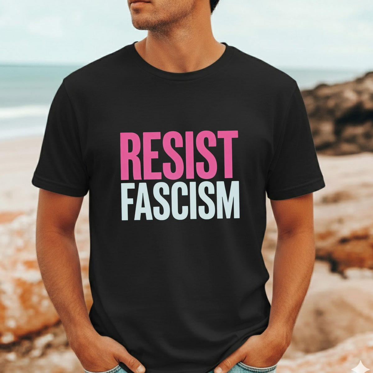A man wearing a black t-shirt with the text 'RESIST FASCISM' printed on the chest in pink and white, set against a blurred beach background.