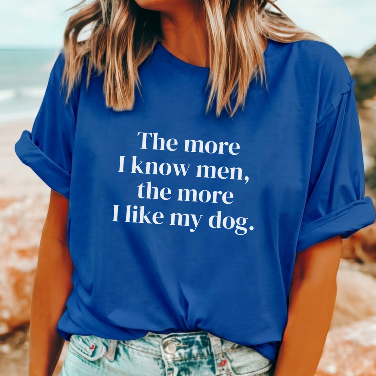 A woman wearing a royal blue t-shirt with the white printed text 'The more I know men, the more I like my dog.' on the front, paired with light-wash denim jeans, set against a blurred beach background.