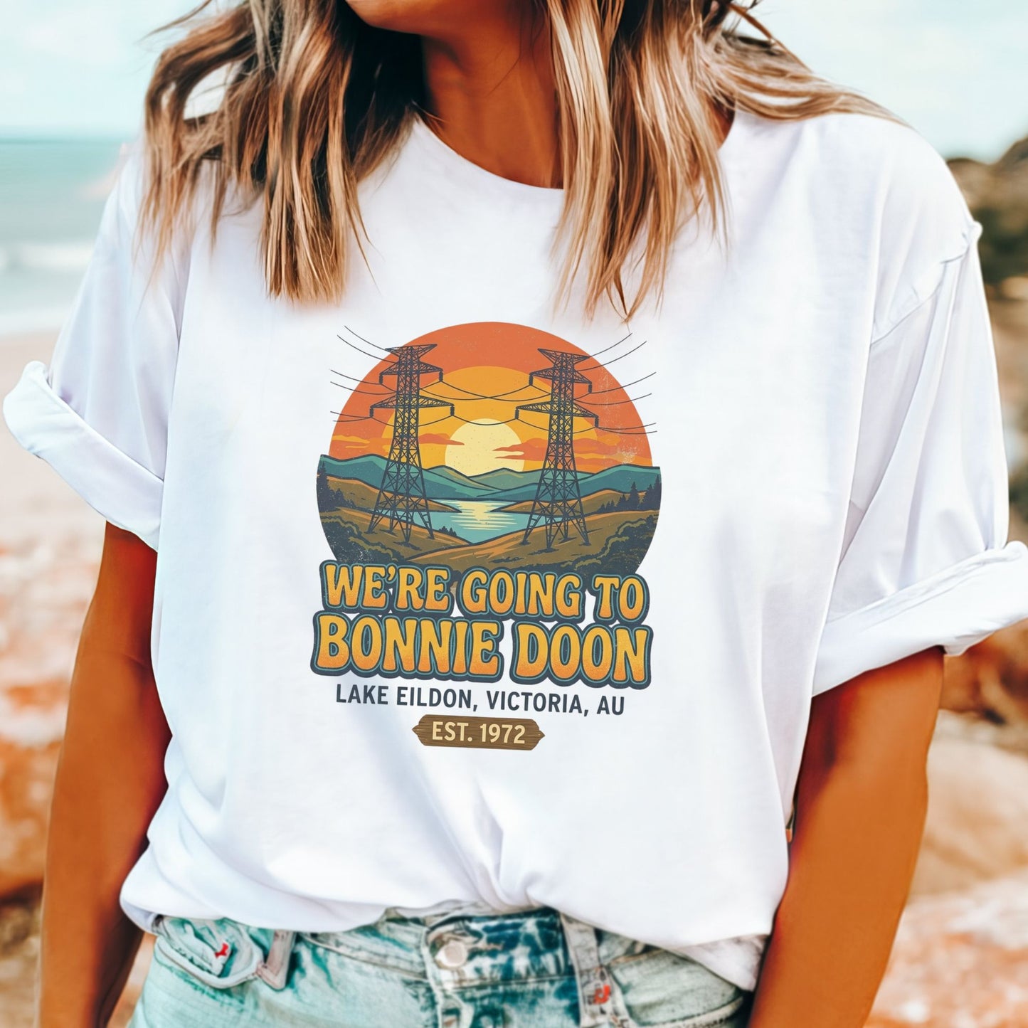 A woman wearing a white graphic t-shirt with a retro-style design that reads 'WE'RE GOING TO BONNIE DOON LAKE EILDON, VICTORIA, AU EST. 1972' with an illustration of power lines and a sunset, paired with light-wash denim jeans.